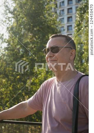 Portrait of smiling adult man wearing sunglasses and pink t-shirt at summer city public park. Outdoors. Vertical format. Close up. 124458470