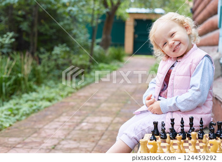 toddler girl smiles at the camera while sitting next to chess on the street in the garden. 124459292