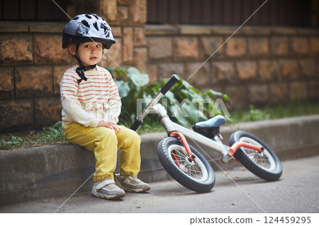 girl in a helmet is sitting on the curb with a running bike lying next to her. girl in a helmet is sitting on the curb with a running bike lying next to her. 124459295