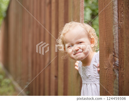 Funny kid in light clothes looks out of a hole in the fence. Summer sunny day. 124459306