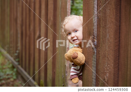 A little curly-haired girl looks out of a hole in the wooden fence. Holds a teddy bear toy. White dress. Summer day. A little curly-haired girl looks out of a hole in the wooden fence. Holds a teddy bear toy. White dress. Summer day. 124459308