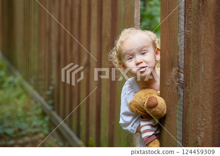 girl with a teddy bear in her hands looks out through a hole in a wooden fence. Copy space. girl with a teddy bear in her hands looks out through a hole in a wooden fence. Copy space. 124459309
