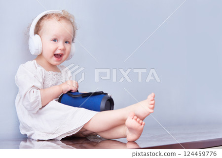 Little cute girl with a music speaker in headphones on her curly head. White dress and light back. 124459776
