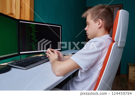 A computer programmer student sits at a computer in his room at a desk on a computer chair. 124459784