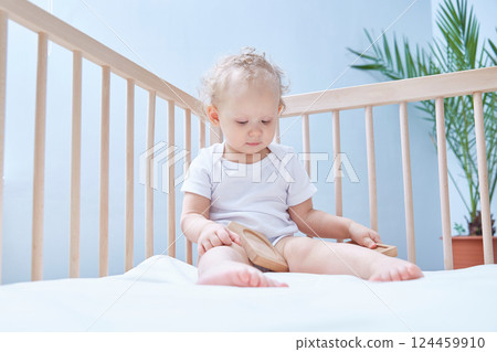 one-year-old baby is sitting in his crib in white clothes in a white room and playing a wooden pyramid 124459910