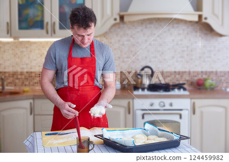 A middle-aged man in a red apron makes dough pies at home in the kitchen. Lifestyle. Copy space 124459982
