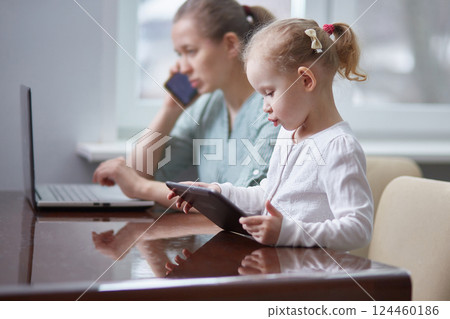 A girl is playing with a tablet. Stressed mother working attending a phone call and taking care of her baby at office A girl is playing with a tablet. Stressed mother working attending a phone call and taking care of her baby at office 124460186