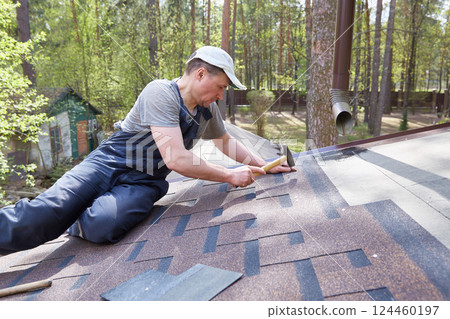 A man mounts soft bitumen shingles with nails and a hammer on the roof of a country house A man mounts soft bitumen shingles with nails and a hammer on the roof of a country house 124460197