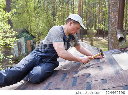 A man mounts soft bitumen shingles with nails and a hammer on the roof of a country house A man mounts soft bitumen shingles with nails and a hammer on the roof of a country house 124460198