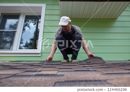 Male worker lays a sheet of soft flexible bitumen shingles on the roof of a country house. Male worker lays a sheet of soft flexible bitumen shingles on the roof of a country house. 124460206