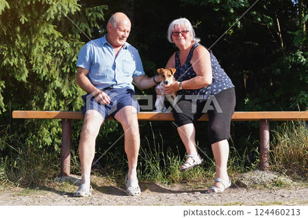 Senior couple sitting on wooden bench with small Jack Russell terrier dog, blurred trees in background Senior couple sitting on wooden bench with small Jack Russell terrier dog, blurred trees in background 124460213