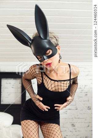 Sexy girl in a short black dress and a rabbit mask from a sex shop poses erotically in a mesh bodysuit 124460314
