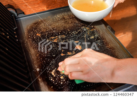 Cleaning electric grill dirty plate, detail on woman hand holding bowl with soup and sponge 124460347