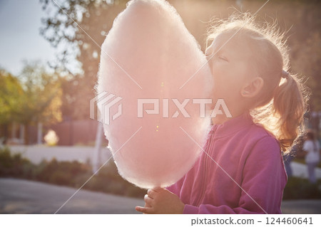 Cute curly-haired little girl eats cotton candy on a sunny summer evening in the park. Close-up. 124460641