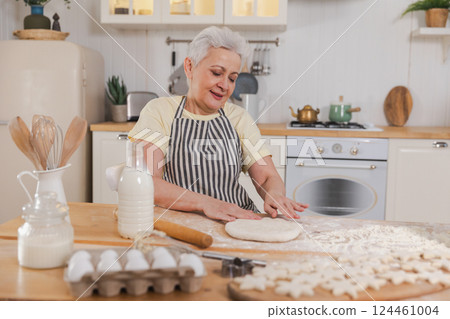 Happy senior woman cooking in kitchen. Stylish older mature gray haired lady grandmother knead dough bake cookies. Old grandma cook homemade food. Household housewife housework concept 124461004
