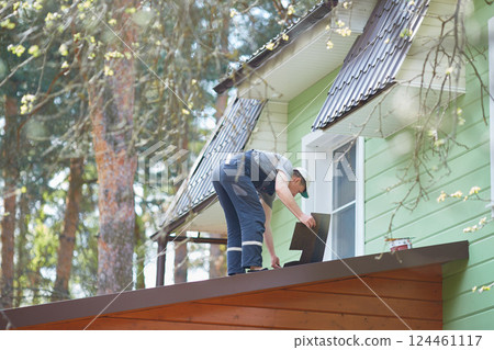 View from the window of the cottage. A person working on the roof installs a soft roof. View from the window of the cottage. A person working on the roof installs a soft roof. 124461117