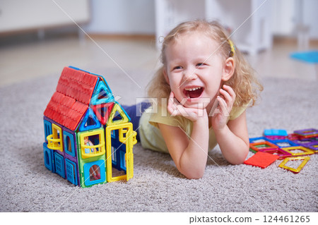 Portrait of a laughing girl on the carpet of the house next to a toy house made of magnets. Portrait of a laughing girl on the carpet of the house next to a toy house made of magnets. 124461265