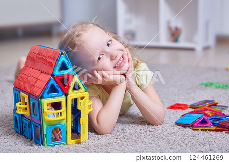 Cute curly girl next to a house made of a magnetic constructor at home on the carpet in the children's room. Cute curly girl next to a house made of a magnetic constructor at home on the carpet in the children's room. 124461269