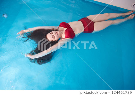 A young woman enjoys relaxing in a swimming pool or a bath with salt water. A brunette with loose hair is relaxing in a spa salon. Top view. Copy space. A young woman enjoys relaxing in a swimming pool or a bath with salt water. A brunette with loose hair is relaxing in a spa salon. Top view. Copy space. 124461789
