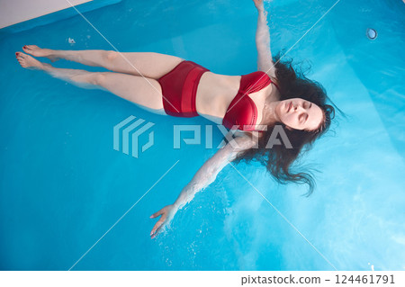 A young woman enjoys relaxing in a swimming pool or a bath with salt water. A brunette with loose hair is relaxing in a spa salon. Top view. Copy space. A young woman enjoys relaxing in a swimming pool or a bath with salt water. A brunette with loose hair is relaxing in a spa salon. Top view. Copy space. 124461791