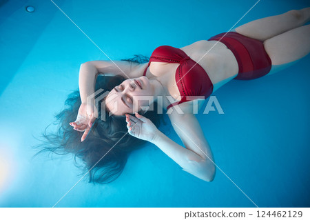 A young woman is swimming relaxed in a small pool, the blue background is visible through the clear water illuminated by light, dark hair, burgundy swimsuit, a day of relaxation at the spa. A young woman is swimming relaxed in a small pool, the blue background is visible through the clear water illuminated by light, dark hair, burgundy swimsuit, a day of relaxation at the spa. 124462129