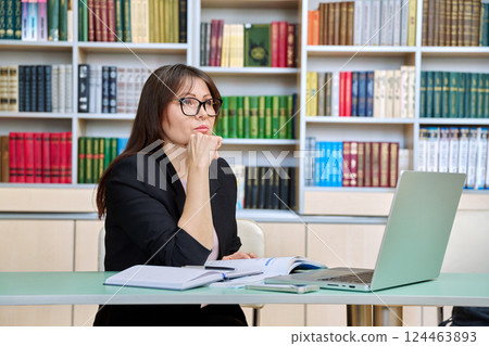 Middle aged female teacher working inside library office with laptop Middle aged female teacher working inside library office with laptop 124463893