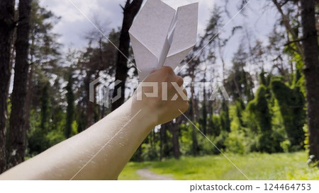 Female hand playing with paper airplane at summer forest. Girl walking with toy plane at woodland on sunny day. Young woman having fun at nature. Concept of leisure outdoor. Close up 124464753