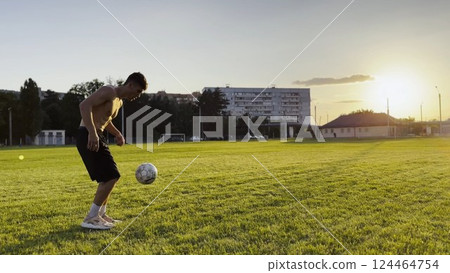 Young man juggling soccer ball on stadium at sunset. Professional footballer kicking ball at green field. Sportsman practicing tricks at meadow with sunlight at background. Freestyle football 124464754