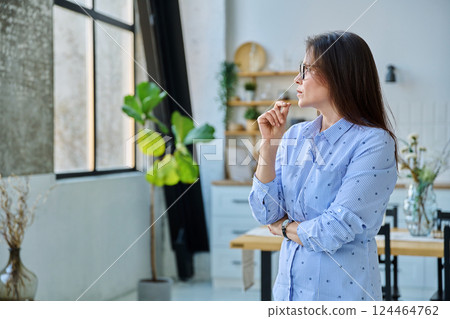 Portrait of serious middle aged woman looking out window in home Portrait of serious middle aged woman looking out window in home 124464762