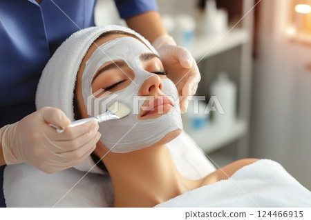 A woman lying with her eyes closed while a beautician applies a thick facial mask with a brush 124466915