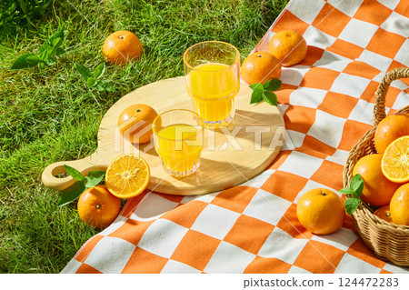 Glasses of juice on wooden cutting board with handles, surrounded by fresh tangerines and leaves. Carpet spread on turf with upright blades of grass. Bamboo basket containing rough-skinned fruits 124472283