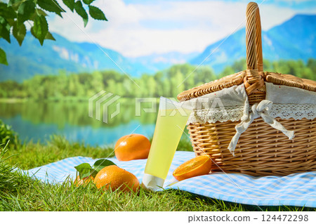 Unlabeled tube of yellow liquid cream, leaning diagonally against a hand-woven wicker basket. Citrus with vibrant hues placed around on a fabric rug. Natural setting, concept of a picnic in the park. 124472298