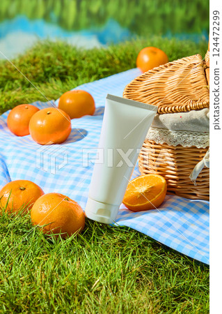 Oranges placed on checkered carpet and on lawn with upright grass blades. White cream tube placed between oranges and handwoven wicker basket. vertical shot for advertising, outdoor picnic concept 124472299