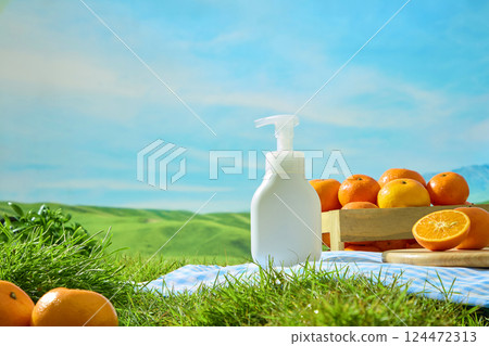 Plain white cosmetic pump bottle placed on checkered carpet and green grass. Fresh oranges placed in wooden crate. Green field landscape with outdoor nature concept. Bottle mockup for design. 124472313