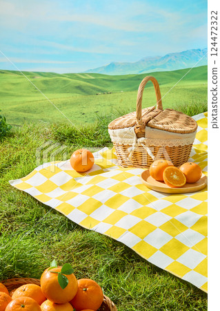 Citrus Burst Orange stacked on top of each other with green stems. Yellow and white checkered picnic mat spread out on a prairie. Plate of fresh oranges and wicker basket filled with oranges 124472322