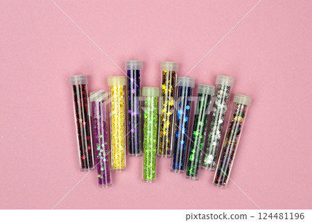An assortment of bright, shiny sequins in small jars scattered across a pink background, catching the light perfectly. 124481196