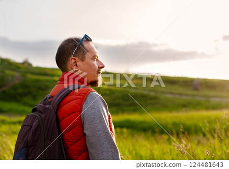 Man hiker wearing backpack and red vest stands outdoors, enjoying serene nature walk with the sun setting in the background. He appears relaxed as he takes in the peaceful scenery, copy space 124481643