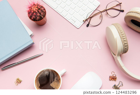 Pastel pink desk with coffee, headphones, and office supplies framing empty space 124483296