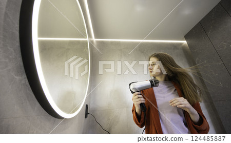 Young woman in a red robe drying her long hair with a hairdryer in a modern bathroom with a glowing round mirror. Young woman in a red robe drying her long hair with a hairdryer in a modern bathroom with a glowing round mirror. 124485887