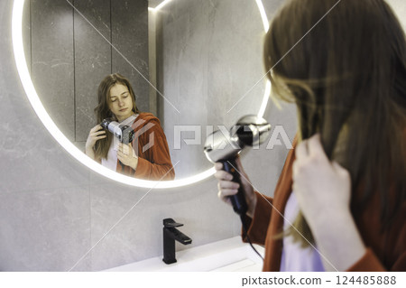 Young woman in a red robe drying her hair with a hairdryer while looking at herself in the round illuminated bathroom mirror. Young woman in a red robe drying her hair with a hairdryer while looking at herself in the round illuminated bathroom mirror. 124485888
