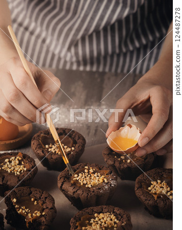 Bakers hands grease the muffins with a brush with egg yolk on a table, close-up, vertical image. Bakers hands grease the muffins with a brush with egg yolk on a table, close-up, vertical image. 124487278