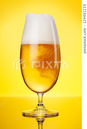 A tall glass of beer with a golden hue and frothy head, on a yellow background 124491219