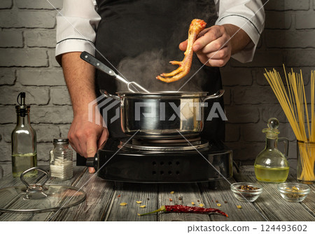 A chef focuses on a pot of simmering broth, carefully adding chicken feet. Fresh ingredients and spices are laid out on a rustic grey wooden table A chef focuses on a pot of simmering broth, carefully adding chicken feet. Fresh ingredients and spices are laid out on a rustic grey wooden table 124493602