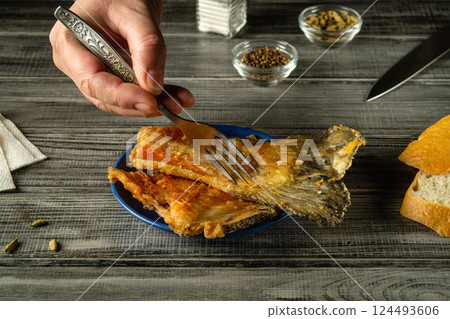 A hand holds a fork poised above a plate of golden fried fish, accompanied by pieces of crusty bread, on a wooden table that adds warmth to the dining experience A hand holds a fork poised above a plate of golden fried fish, accompanied by pieces of crusty bread, on a wooden table that adds warmth to the dining experience 124493606