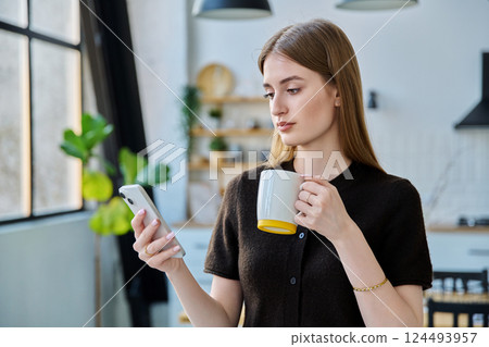 Young woman with cup of drink using smartphone standing at home Young woman with cup of drink using smartphone standing at home 124493957