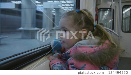 A child in a pink winter coat looks thoughtfully out of the subway car window holding a stuffed animal and watching the cityscape pass by during a quiet ride. Child riding in a subway car. 124494350