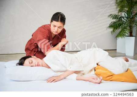 Masseuse making thai yoga massage. Woman therapist doing traditional massage treatment for female patient using elbow. Client lying on mat, meditating, enjoying hand massage in calm atmosphere. Masseuse making thai yoga massage. Woman therapist doing traditional massage treatment for female patient using elbow. Client lying on mat, meditating, enjoying hand massage in calm atmosphere. 124494563