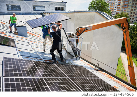Workers building solar panel system on metal rooftop of house with assistance of crane lift. Two men installers carrying photovoltaic solar module outdoors. Renewable energy generation concept. Workers building solar panel system on metal rooftop of house with assistance of crane lift. Two men installers carrying photovoltaic solar module outdoors. Renewable energy generation concept. 124494566