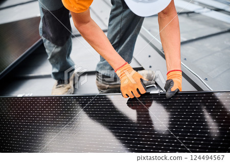 Worker building photovoltaic solar panel system on rooftop of house. Close up of man engineer in gloves installing solar module with help of hex key outdoors. Alternative energy generation concept. 124494567