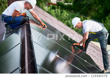 Workers building solar panel system on rooftop of house. Two men installers in helmets installing photovoltaic solar module outdoors. Alternative, green and renewable energy generation concept. Workers building solar panel system on rooftop of house. Two men installers in helmets installing photovoltaic solar module outdoors. Alternative, green and renewable energy generation concept. 124494579